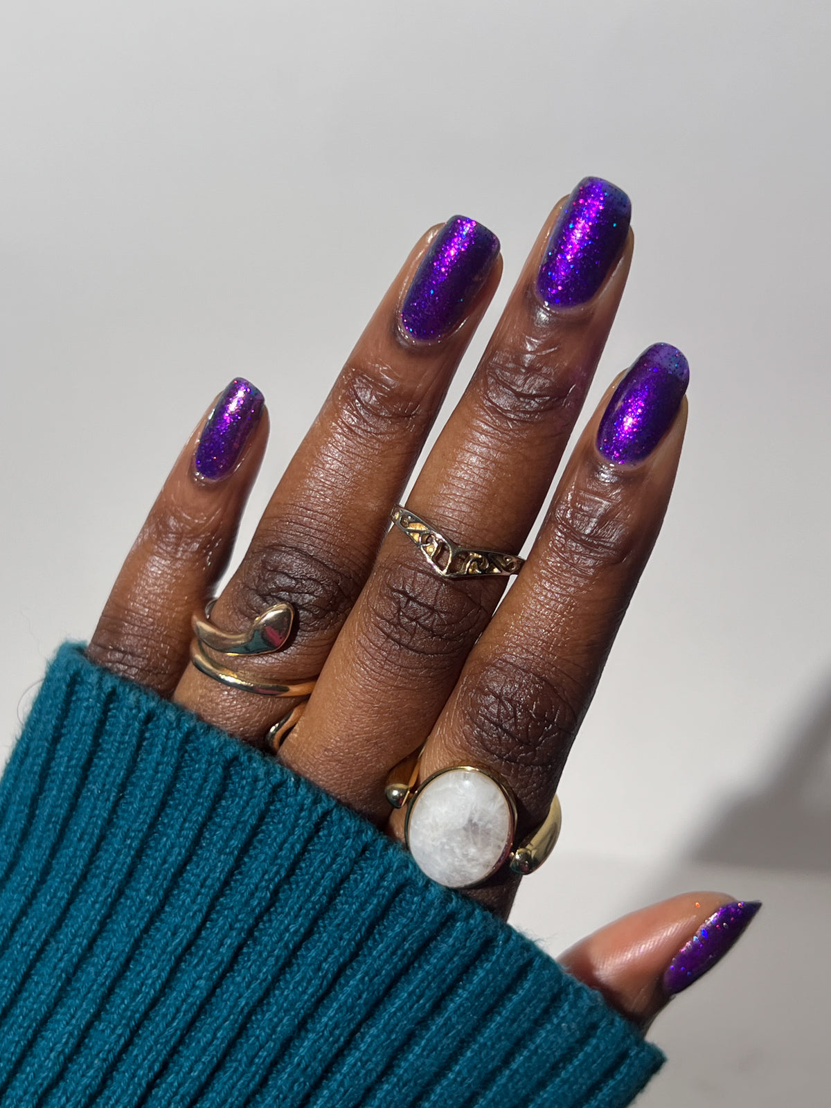 A close-up of a hand with metallic purple nail polish wearing multiple gold rings, including a large ring with a white oval stone, against a plain light background. dark skin tone