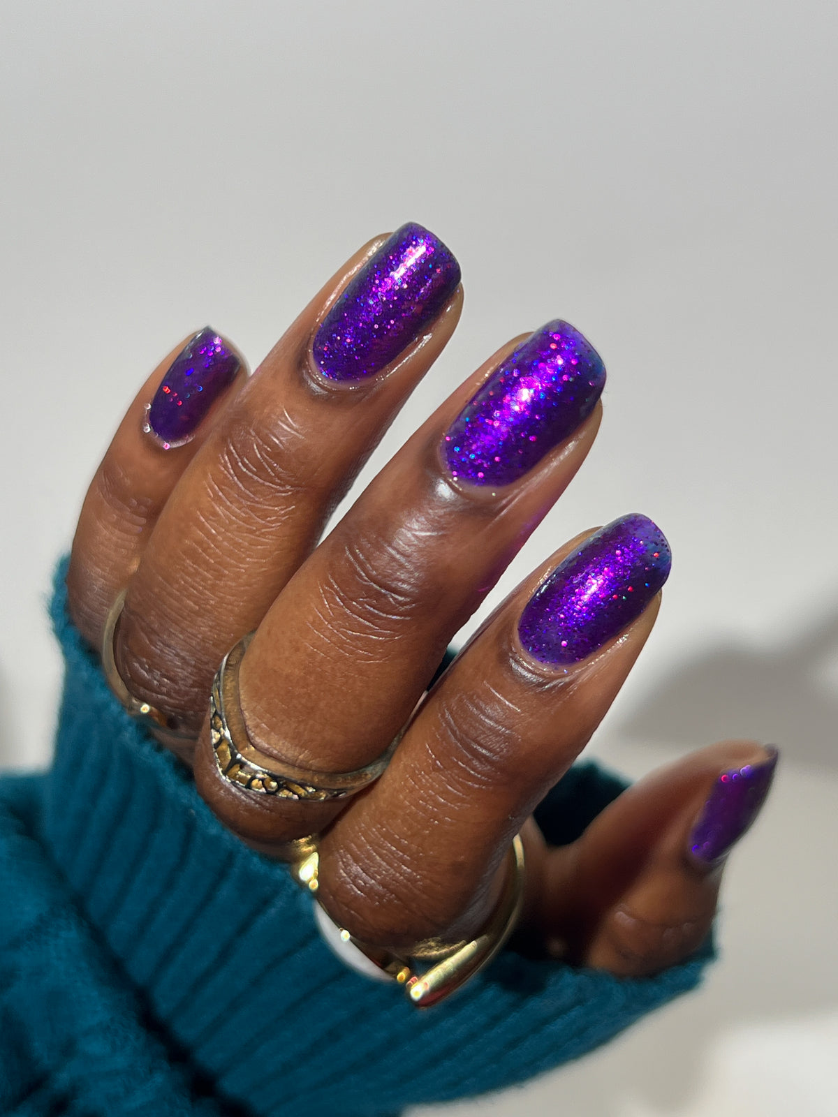 A close-up of a hand with dark skin with manicured nails painted in a sparkling metallic purple polish, wearing gold rings and a teal ribbed sweater cuff against a plain light background.