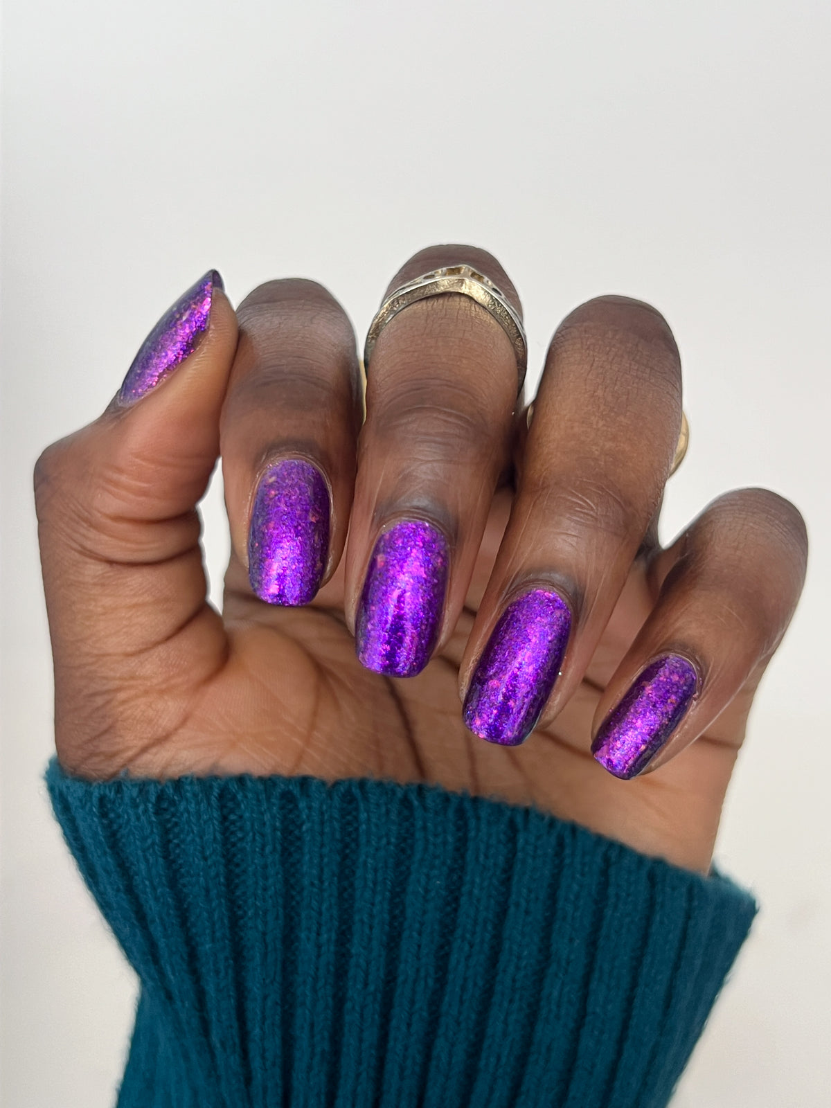 A close-up of a hand with dark skin with manicured nails painted in a sparkling metallic purple polish, wearing a teal ribbed sweater cuff and a gold ring, against a plain light background.