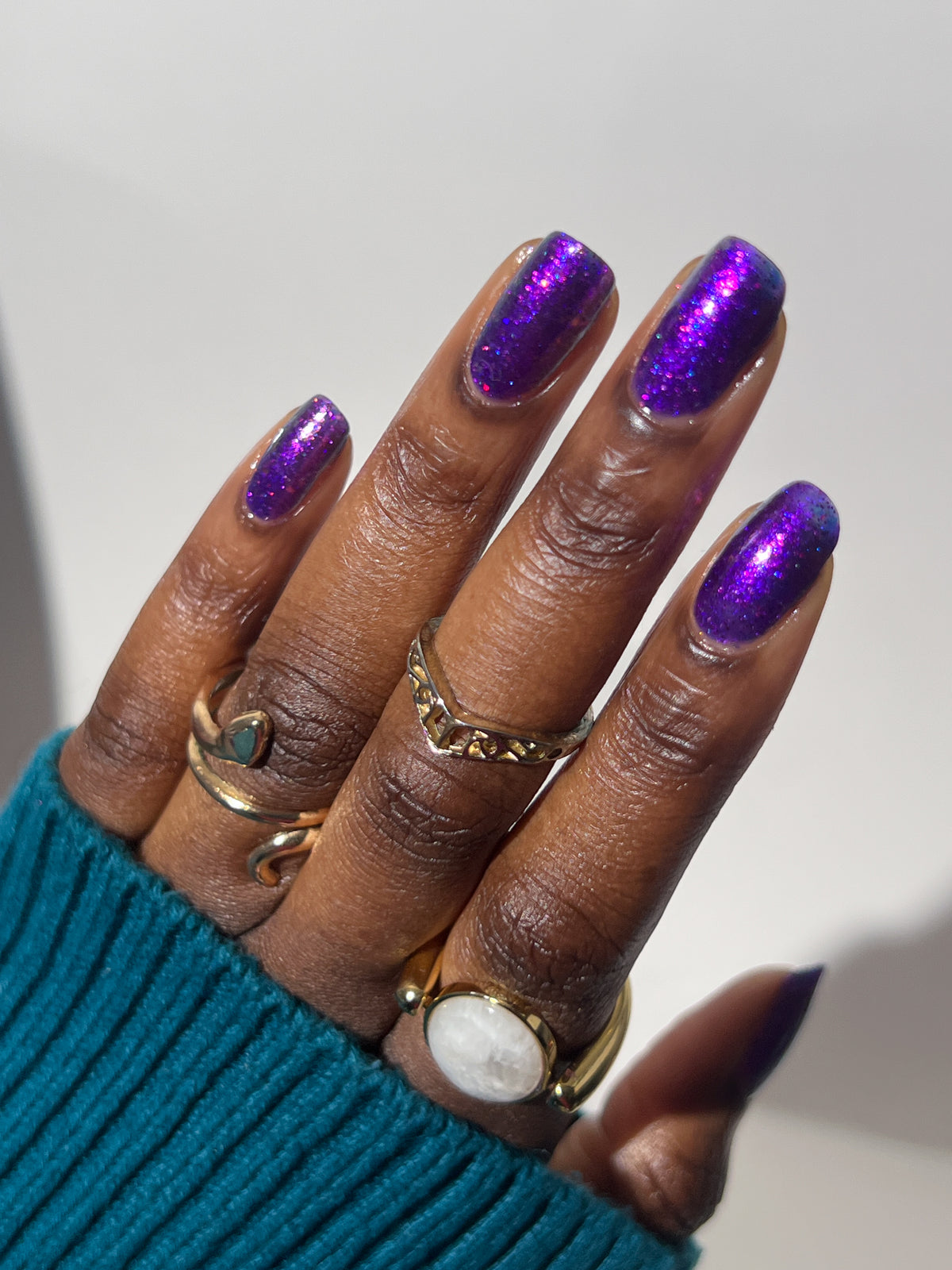 A close-up of a hand with long, glossy purple glitter manicured nails, wearing several gold rings and a teal ribbed sweater cuff, set against a plain light background.