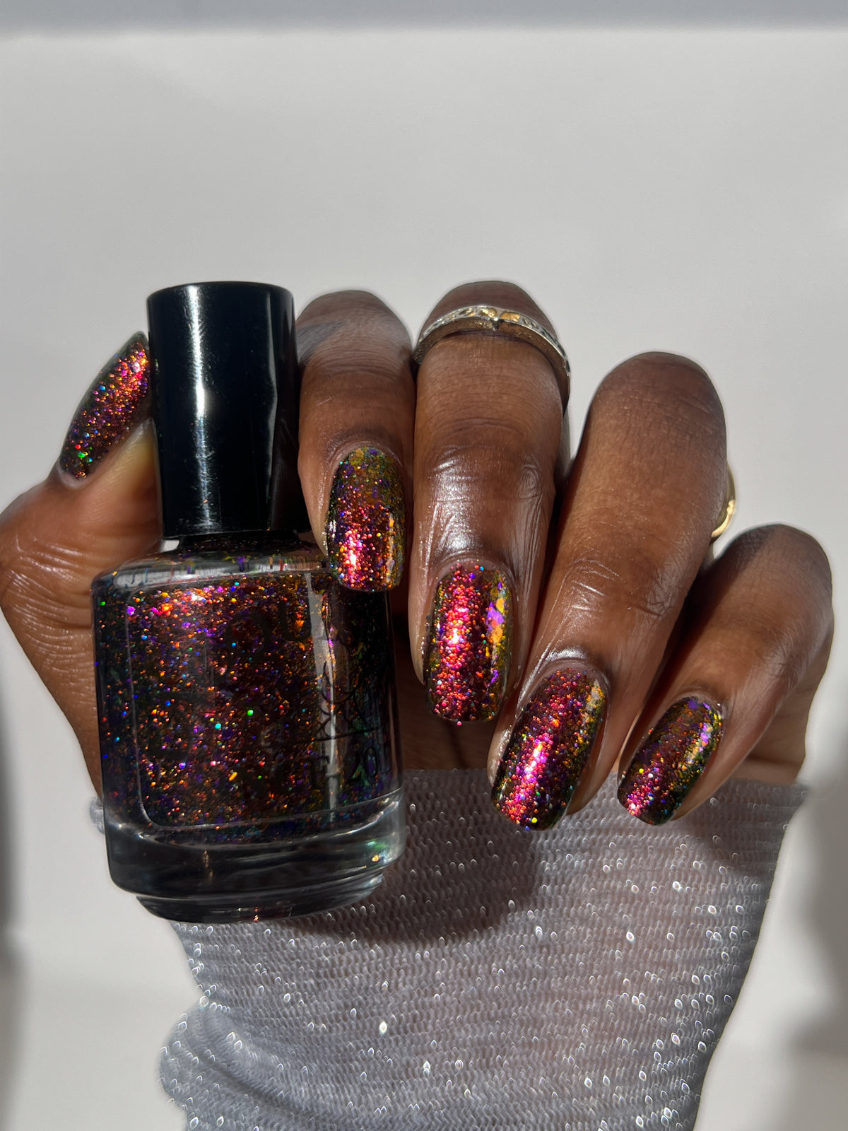 A hand holding a bottle of holographic glitter nail polish, with the nails painted in a matching shimmering pink, purple, and gold shade.