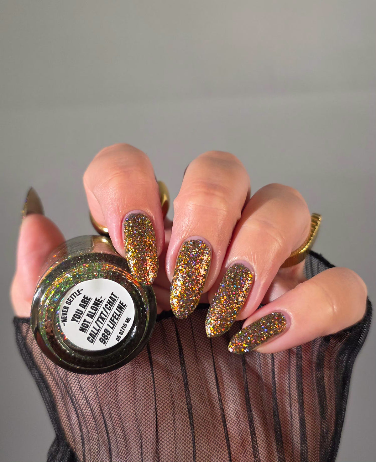nails painted with sparkly glittery polish showing pink, red, green, gold and orange glitters in a darkened base color with green reflective glitter holding bottle that says you are not alone 988 lifeline