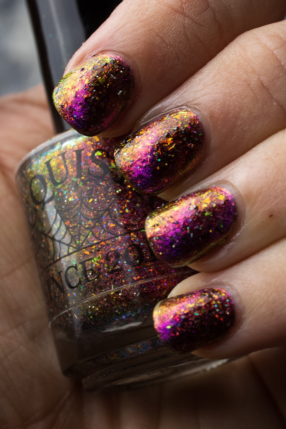 flakie magnetic polish showing red, orange, gold, green flakes, holo glitter, and magnetic effect of fuchsia to purple holding Lacquistry bottle