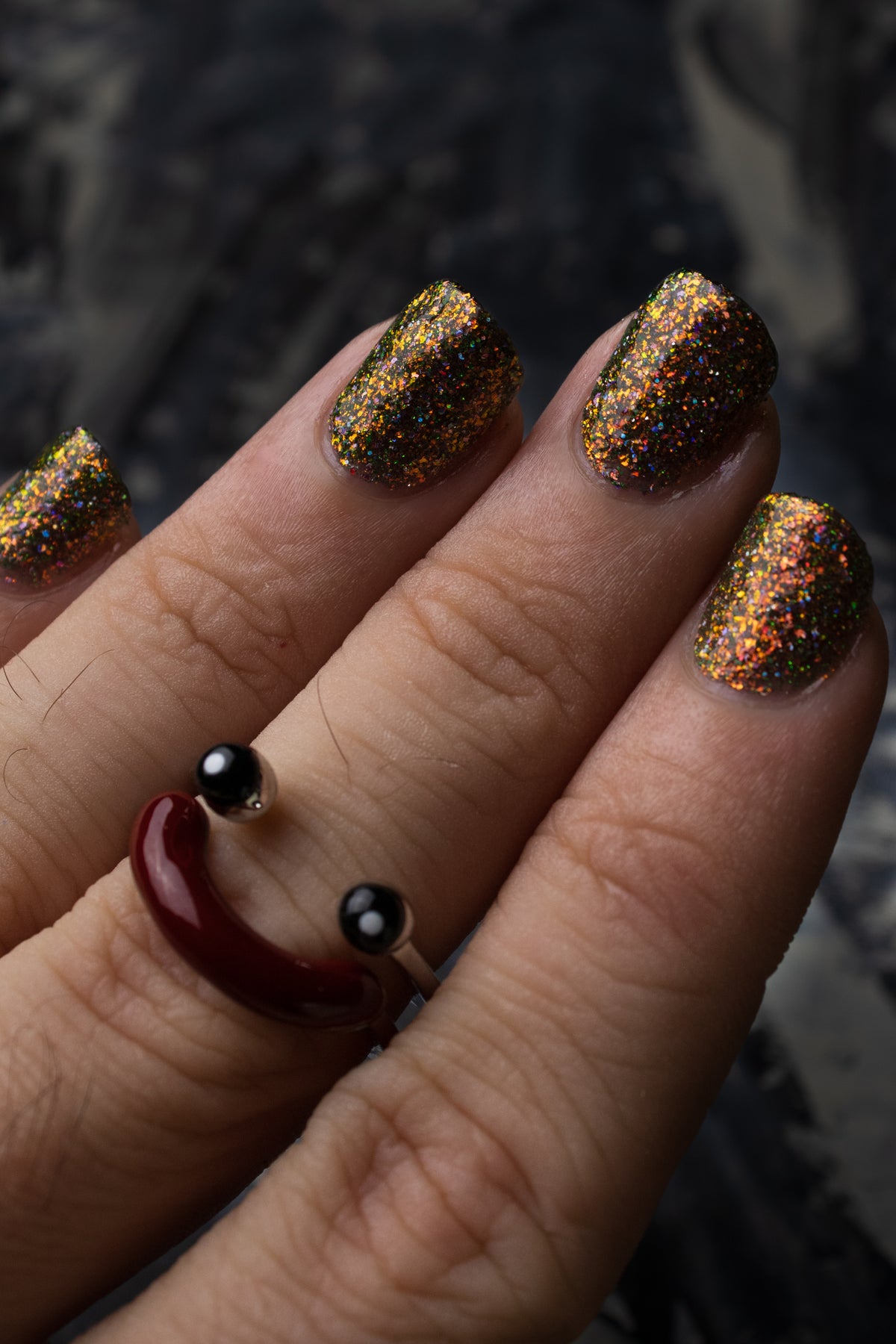 nails painted with sparkly glittery polish showing pink, red, green, gold and orange glitters in a darkened base color with green reflective glitter wearing smiley ring