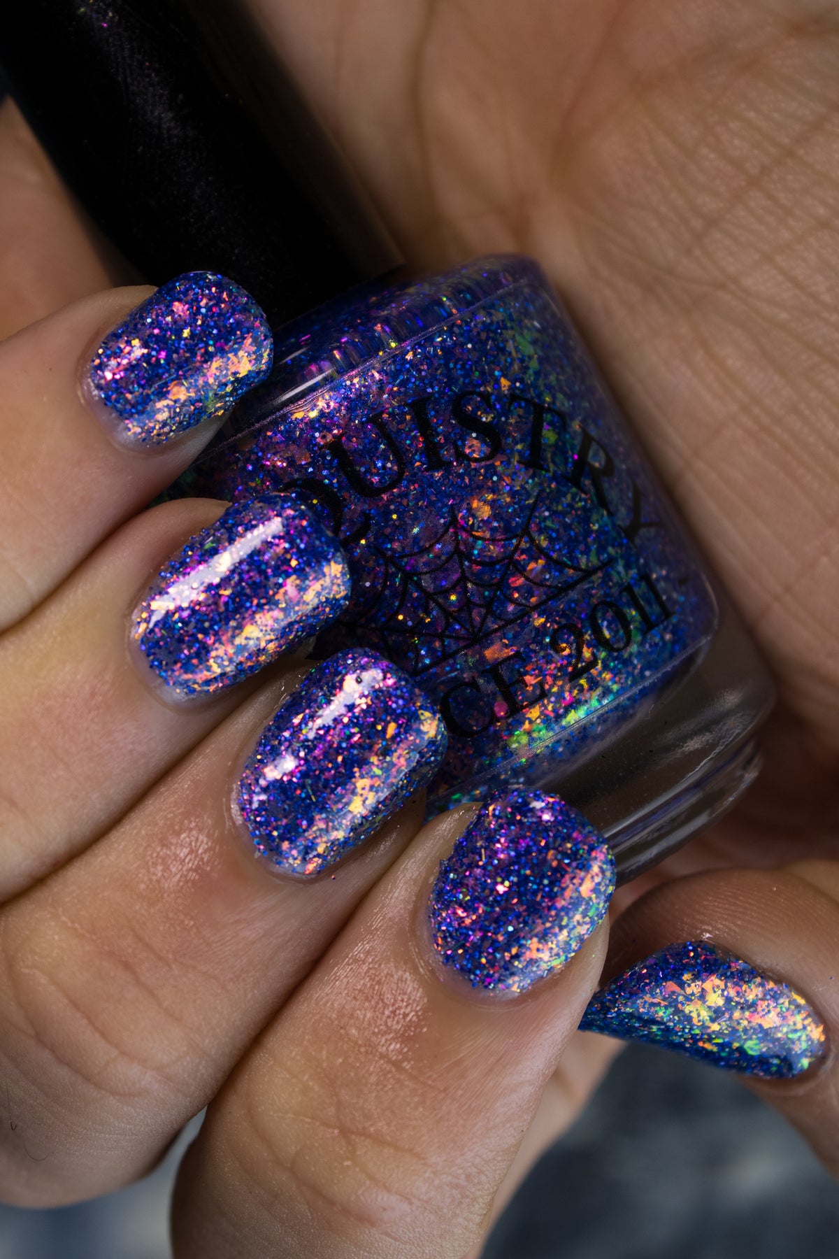 A close-up of a hand holding a bottle of holographic nail polish, with the nails painted in a sparkling blue, purple, and iridescent glitter shade.