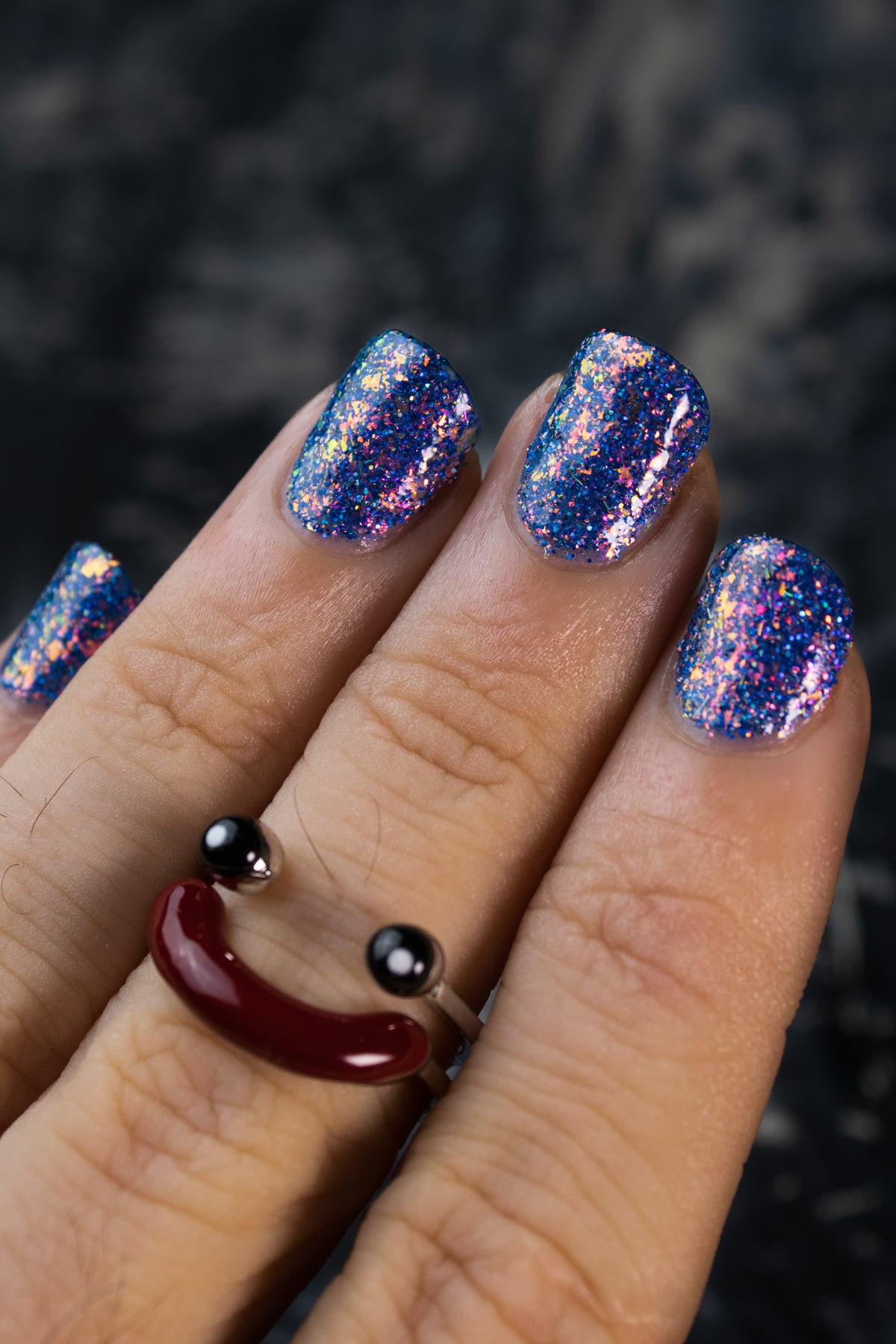 A close-up of a hand with manicured nails painted in a glittery blue and purple holographic nail polish, wearing a smiley face ring on the middle finger against a dark textured background.