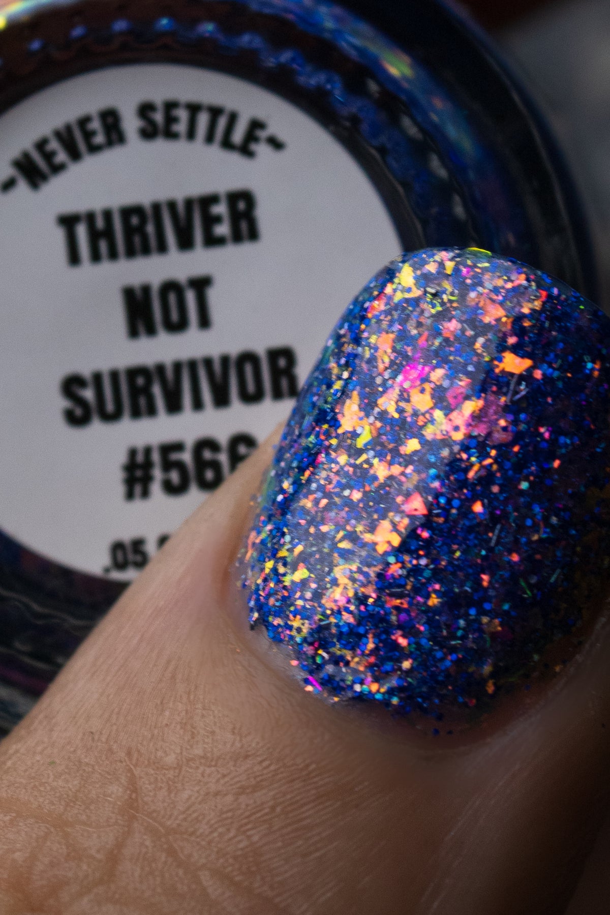 A close-up of a fingernail painted with holographic blue glitter nail polish, with part of the bottle label reading 'NEVER SETTLE THRIVER NOT SURVIVOR #566' visible in the background.