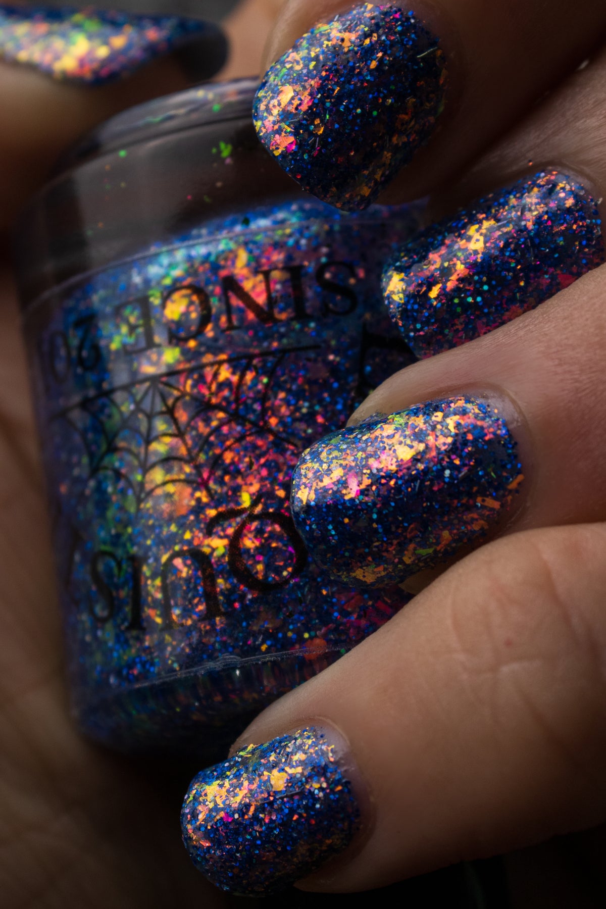 Close-up of a hand with holographic glitter nail polish in a deep blue base, showing iridescent rainbow reflections across the fingernails, with the matching glitter polish bottle held behind the hand.
