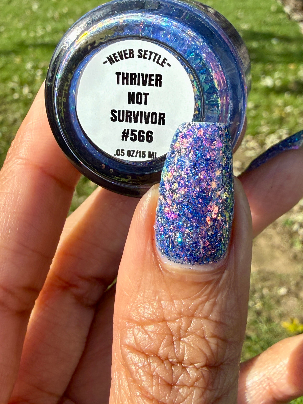 A hand holding a small jar of glitter nail polish outdoors, with the product label reading 'NEVER SETTLE - THRIVER NOT SURVIVOR #566'. The nail polish is a sparkling blue-purple glitter shade shown on the fingernail.