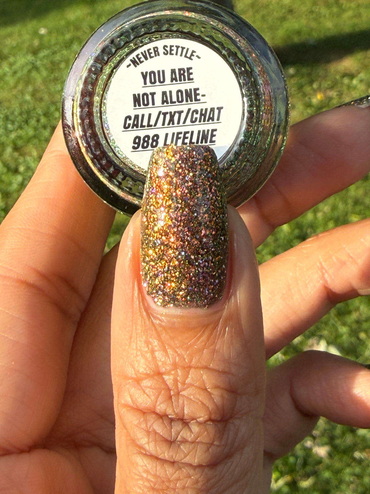 nails painted with sparkly glittery polish showing pink, red, green, gold and orange glitters in a darkened base color with green reflective glitter. bottle says you are not alone 988 lifeline