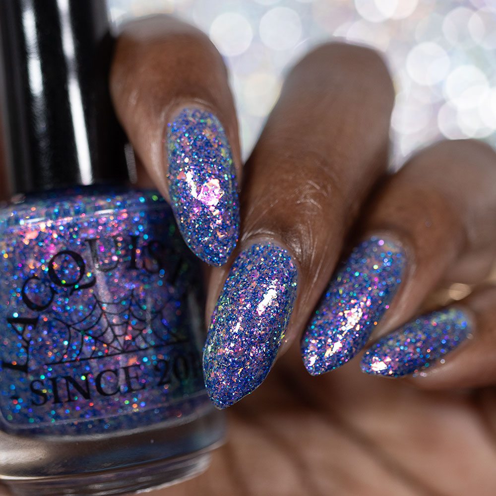 A close-up of a hand with long almond-shaped nails painted in a sparkling holographic blue nail polish, holding a bottle of the same glittery polish. The polish reflects pink, purple, and blue iridescent shimmer.