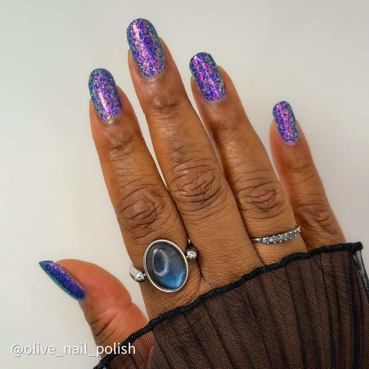 A close-up of a hand showcasing a glittery purple and blue nail polish with a holographic shimmer effect. The hand is wearing silver rings, including one with a large oval blue stone and another with small clear stones, and a sheer black sleeve.