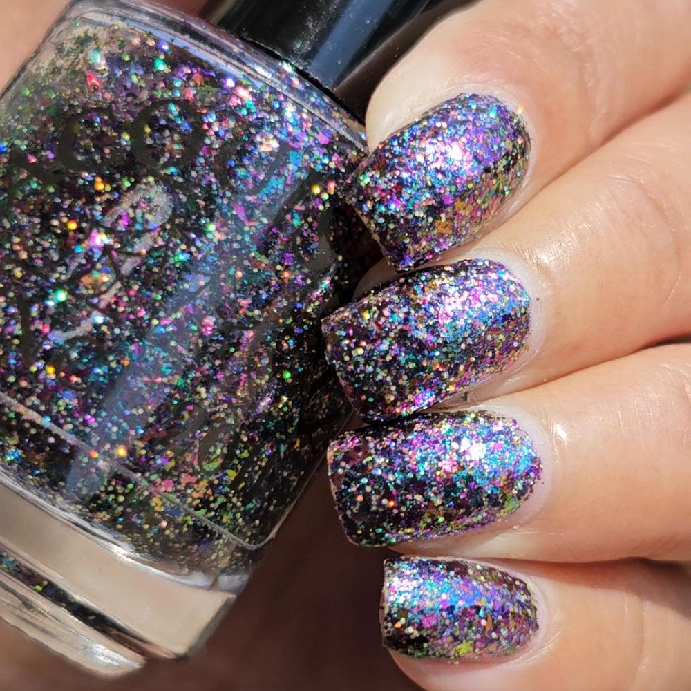 A close-up of a hand with nails painted in a dense, iridescent glitter nail polish, holding a matching nail polish bottle. The polish has a black base covered in holographic, rainbow-colored glitter.