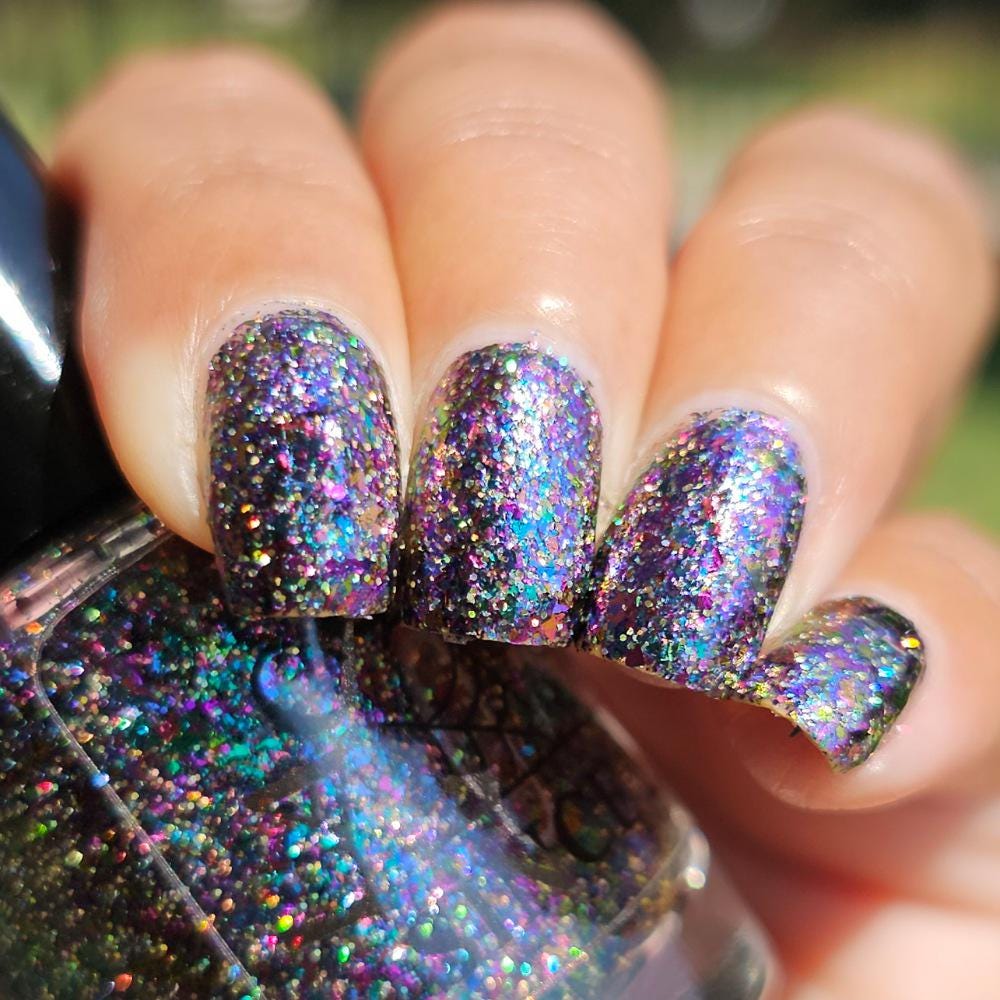 A close-up of a hand with manicured nails painted in a dark glitter nail polish that shifts through purple, blue, green, and pink, with a matching glittery nail polish bottle in the foreground.