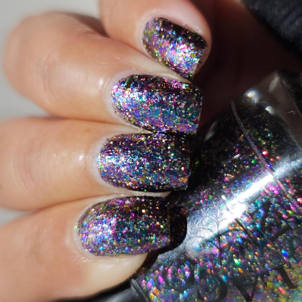 A close-up of a hand with nails painted in a dense, iridescent holographic glitter polish, next to the matching glitter nail polish bottle.