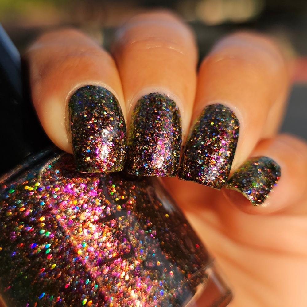 A close-up of a medium skin tone hand with manicured nails painted in a dark multichrome burgundy glitter nail polish that reflects holographic rainbow sparkle, holding a matching glitter nail bottle.
