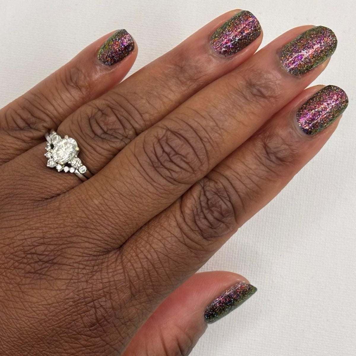 A close-up of a dark skin toned hand with manicured nails painted in a dark multichrome burgundy glitter nail polish that reflects holographic rainbow sparkle, wearing a diamond ring