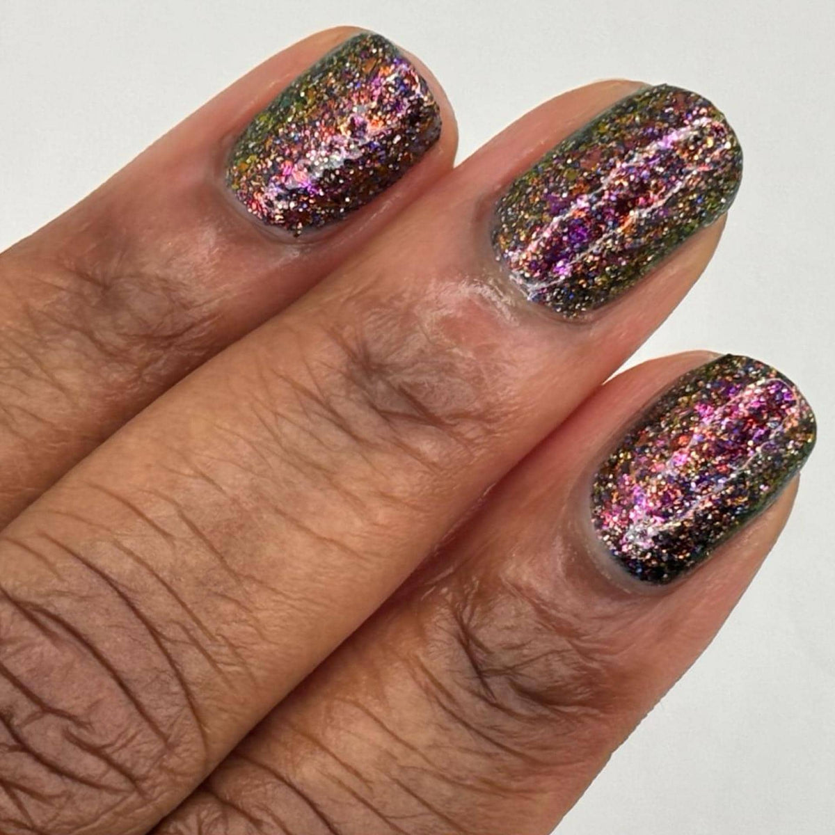 A close-up of a dark skin tone hand with manicured nails painted in a dark multichrome burgundy glitter nail polish that reflects holographic rainbow sparkle