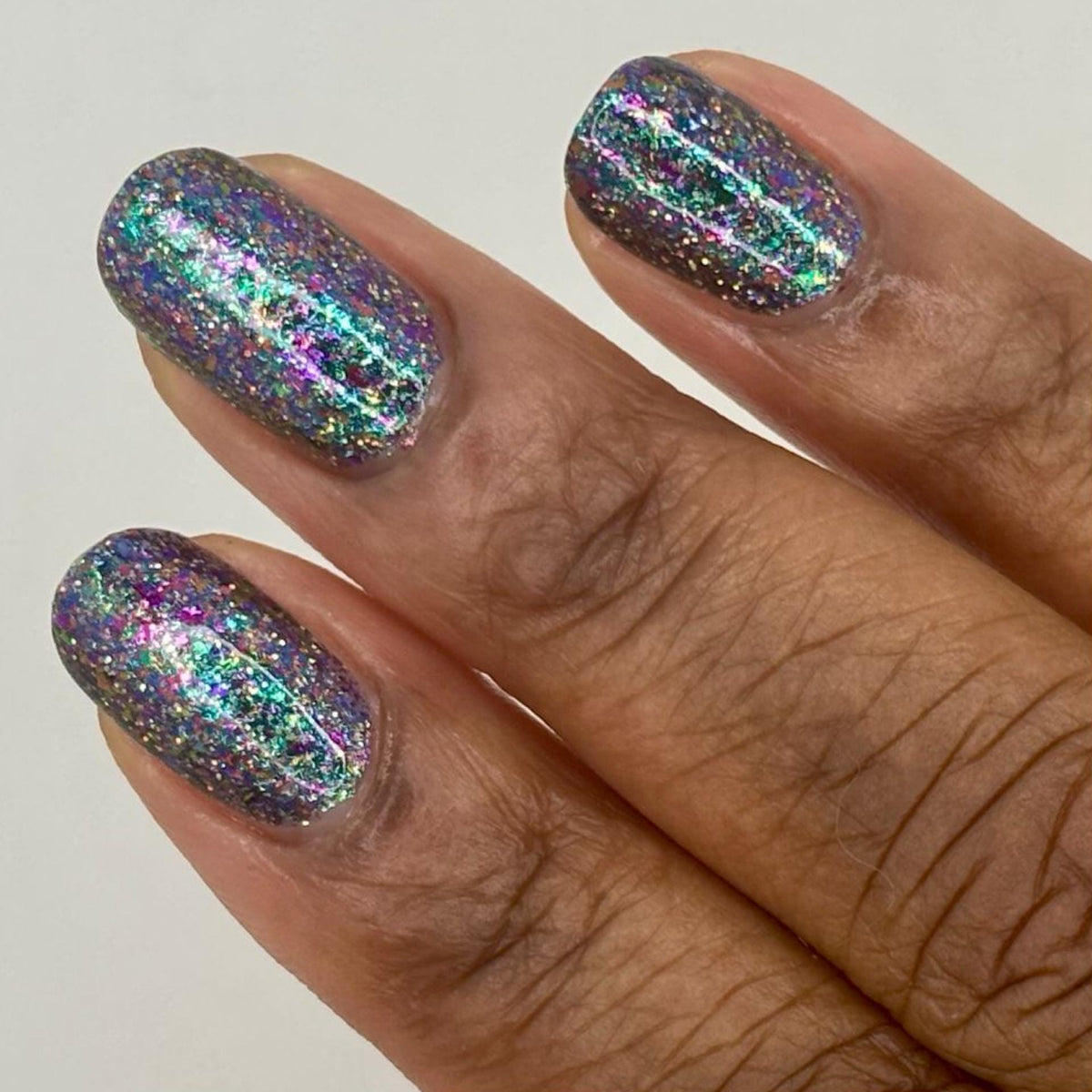 A close-up of a hand with nails painted in a dark holographic glitter nail polish, holding a matching nail polish bottle. The polish reflects rainbow iridescent glitter.