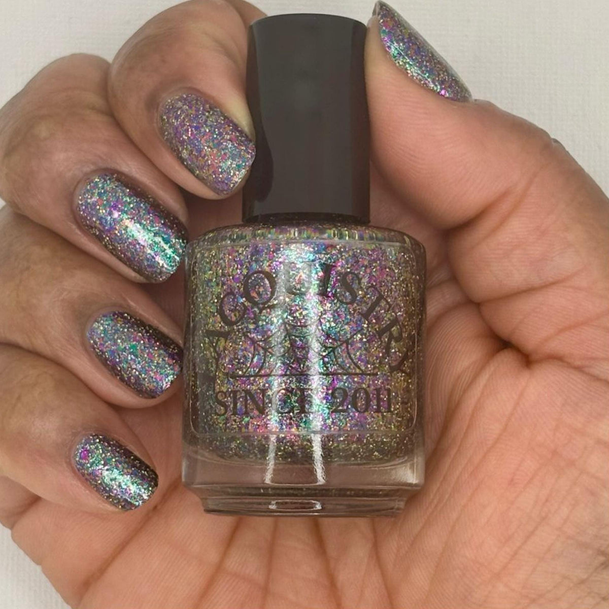 A close-up of a hand with nails painted in a dark holographic glitter nail polish, holding a matching nail polish bottle. The polish reflects rainbow iridescent glitter.