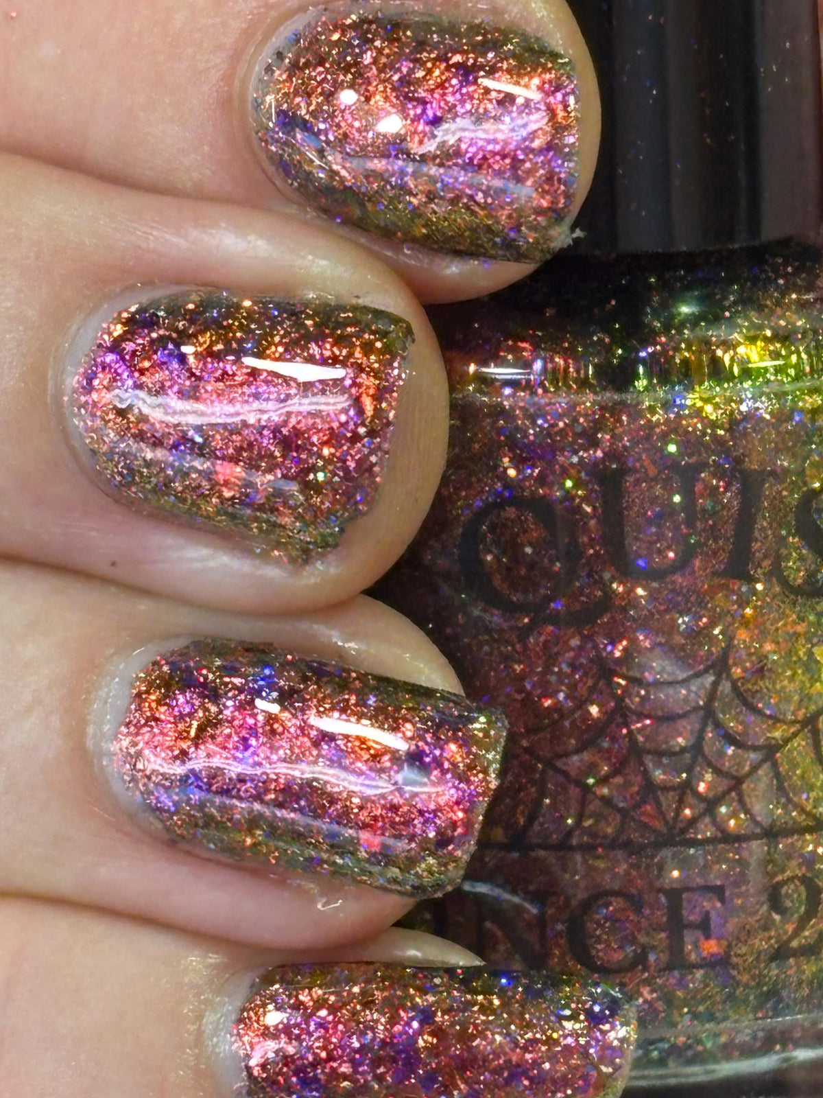 Close-up of a hand with manicured nails painted in a holographic glitter nail polish showing pink, purple, orange, and green iridescent reflections.