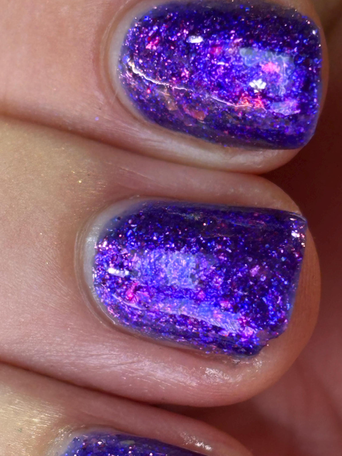 A close-up of fingernails painted with a chunky, highly reflective purple glitter nail polish that sparkles with pink and blue tones.