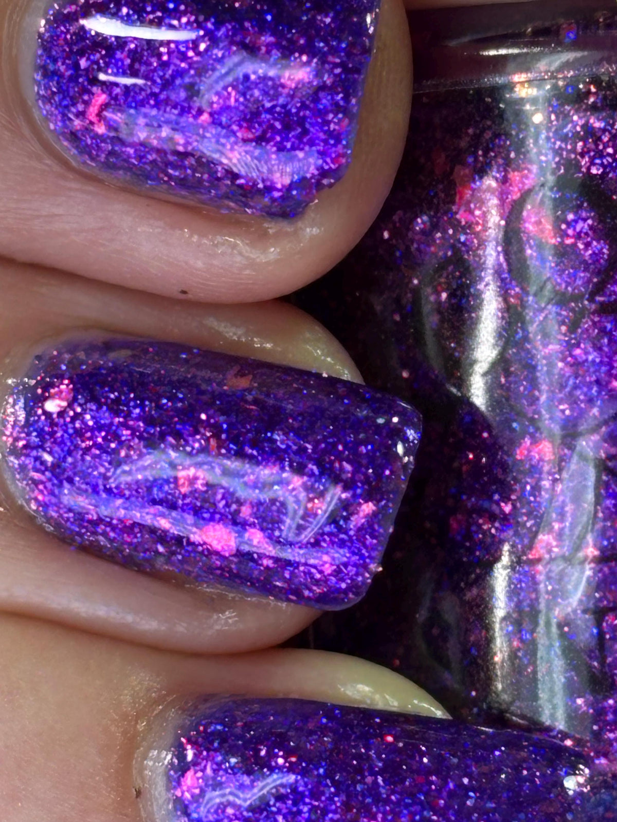 Close-up of fingernails painted with a flakey, glittery purple nail polish featuring bright pink and blue reflective flecks, with a bottle of the same polish partially visible on the right.