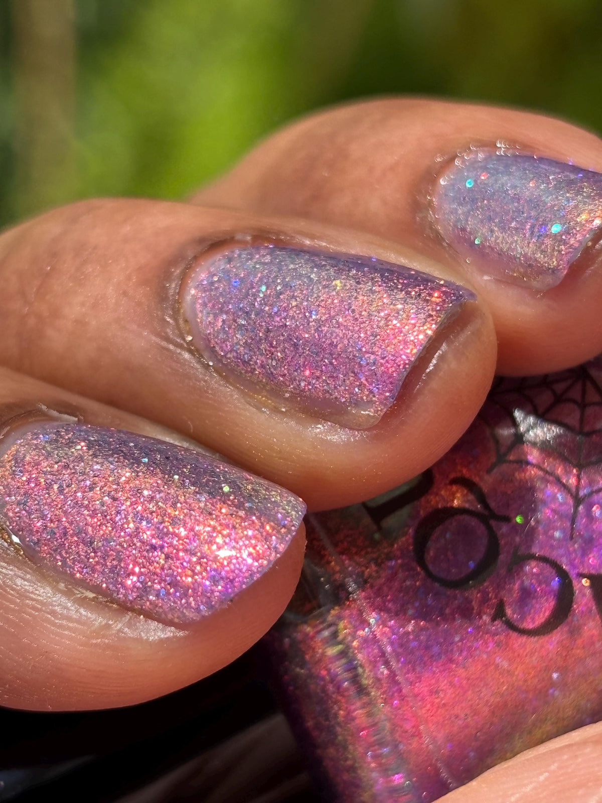 darker skin toned hand with short nails wearing metallic purple pink sparkle nail polish holding matching bottle of light purple nail polish