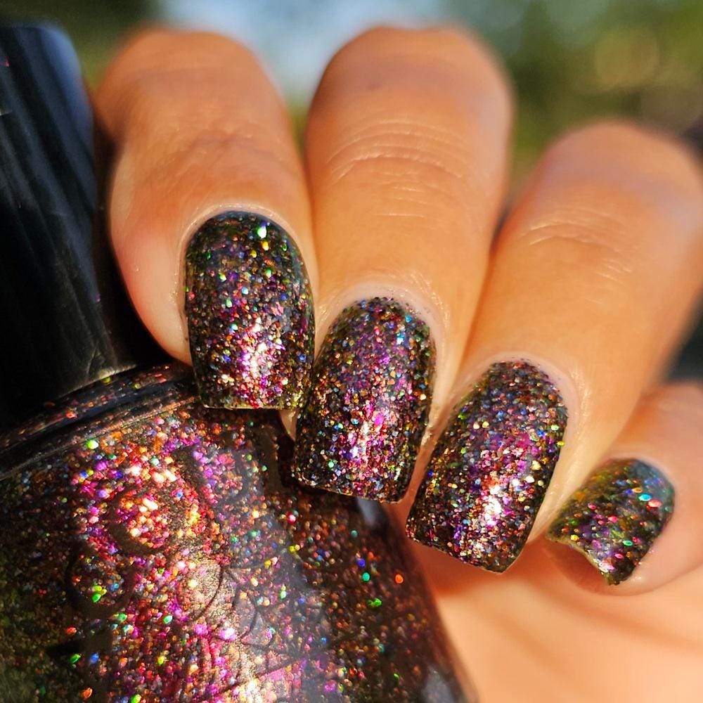 A close-up of a medium skin tone hand with manicured nails painted in a dark multichrome burgundy glitter nail polish that reflects holographic rainbow sparkle, holding a matching glitter nail bottle.
