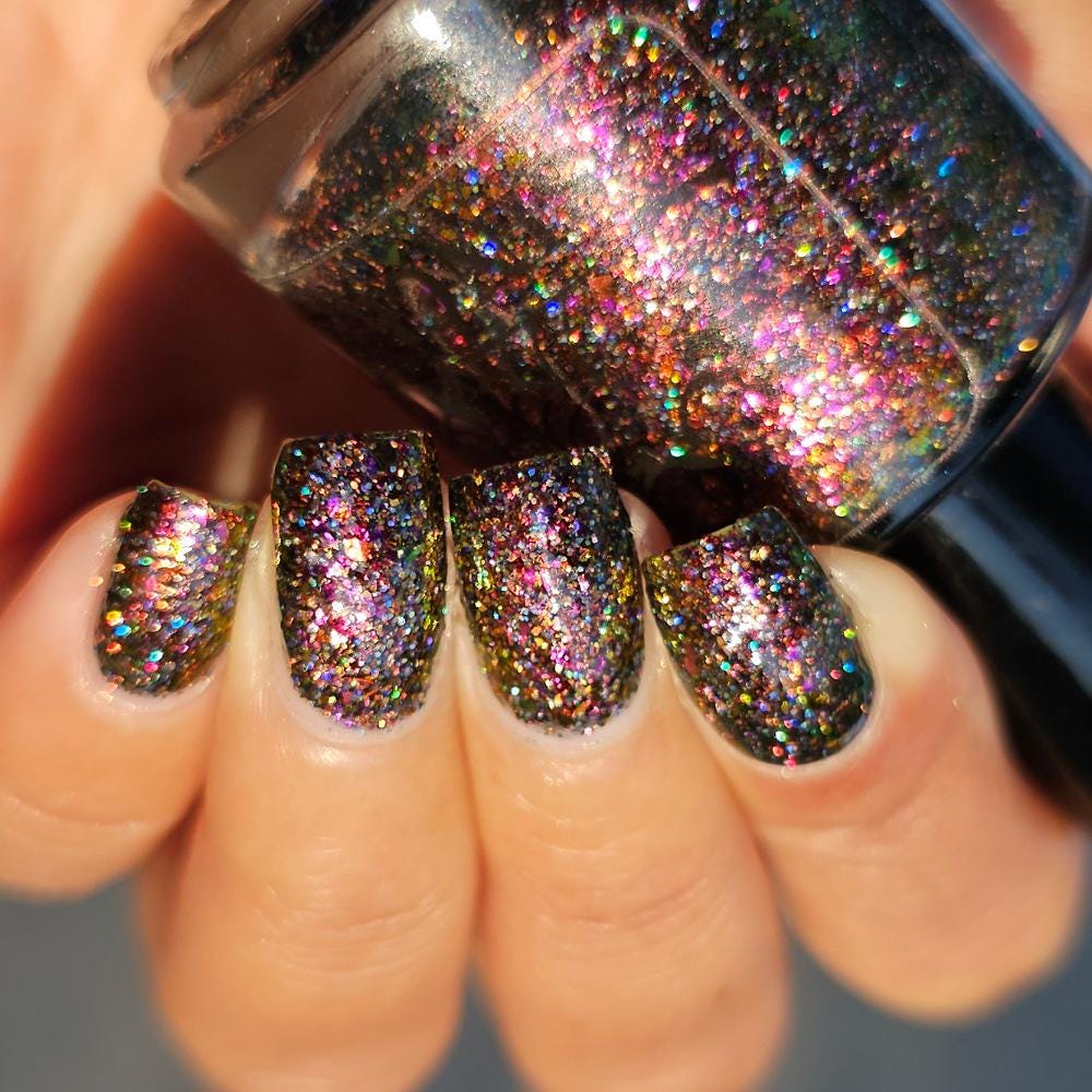 A close-up of a medium skin tone hand with manicured nails painted in a dark multichrome burgundy glitter nail polish that reflects holographic rainbow sparkle, holding a matching glitter nail bottle.