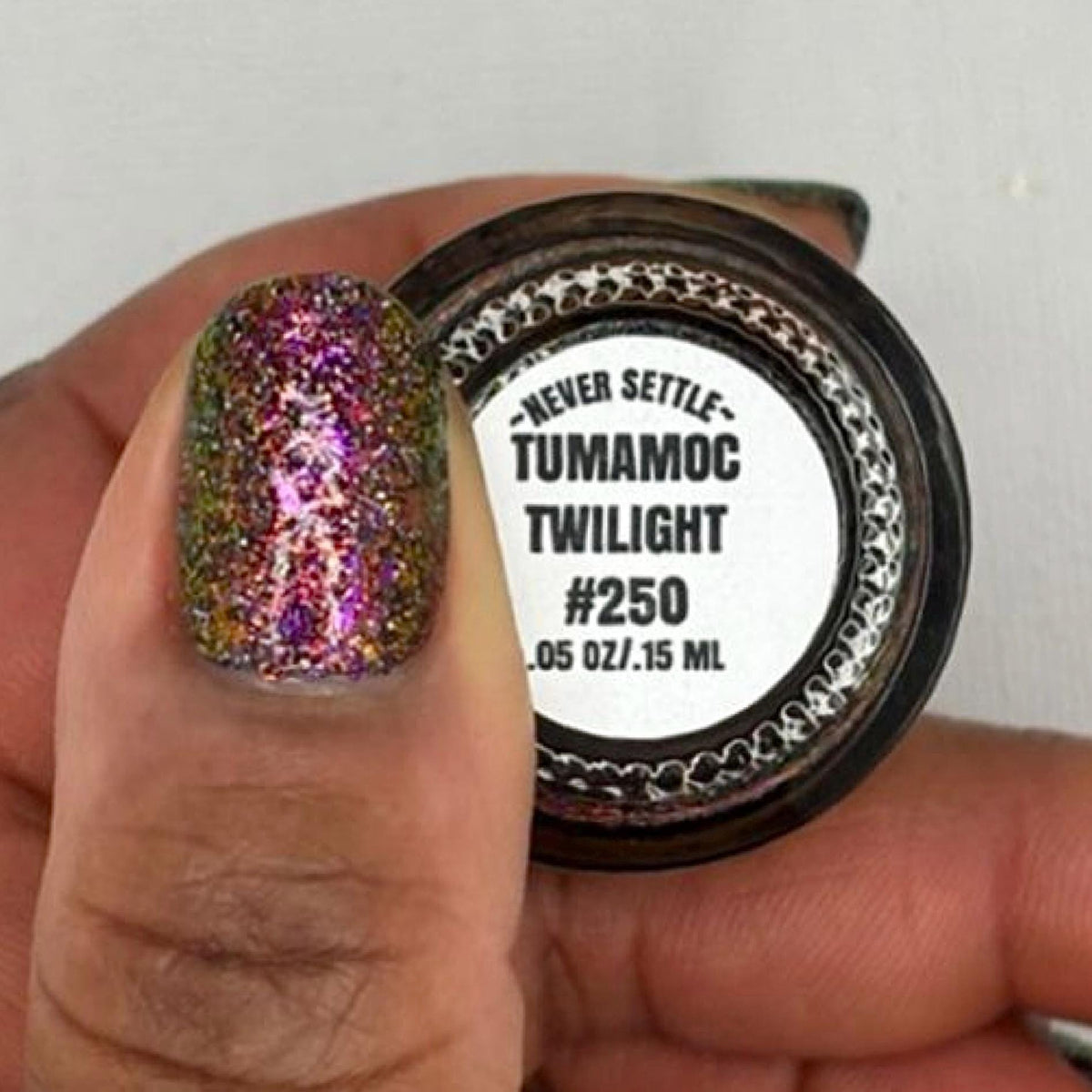close-up of a dark skin tone thumb with manicured nails painted in a dark multichrome burgundy glitter nail polish that reflects holographic rainbow sparkle, holding a nail polish bottle showing the bottom that says "never settle tumamoc twilight #250 .05 oz 15ml"