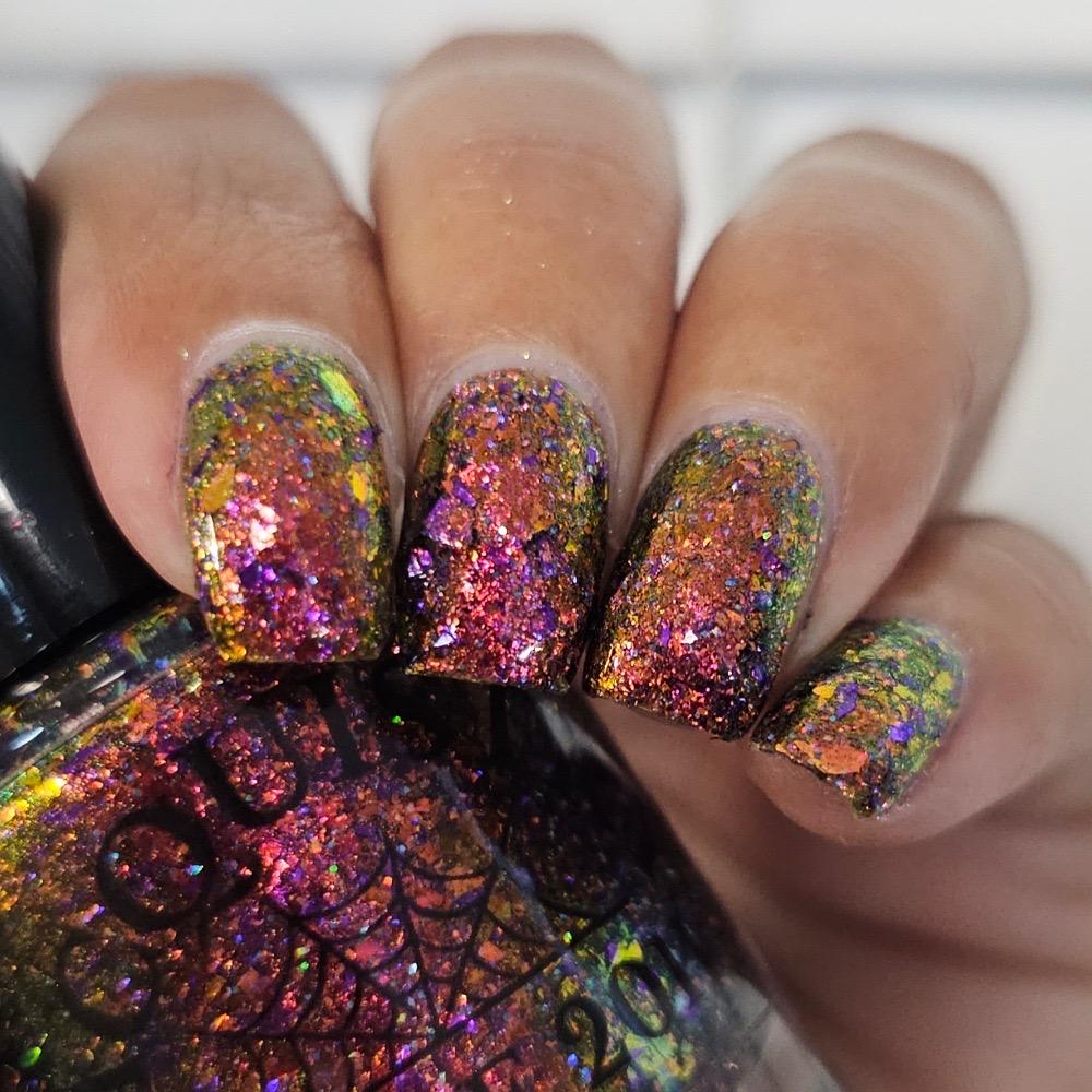 Close-up of a hand with manicured nails painted in a holographic glitter nail polish showing pink, purple, orange, and green iridescent reflections.