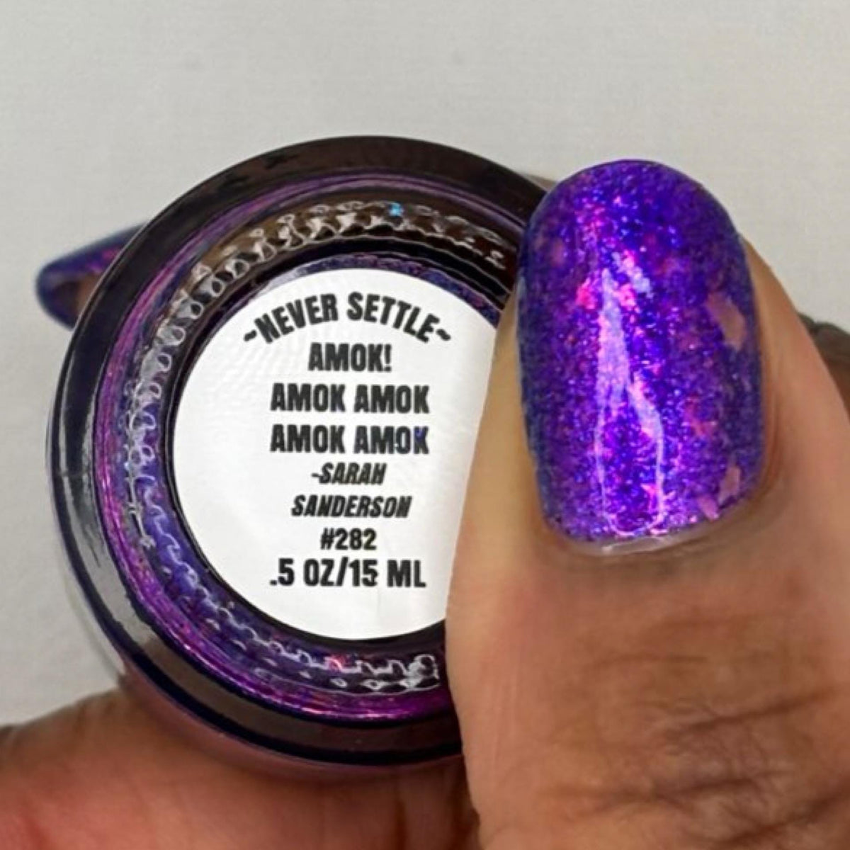 A close-up of a nail polish bottle with its lid held by a hand with dark skin showing the applied color. The nail polish is a sparkling purple with strong pink and blue iridescent shimmer, labeled 'AMOK! AMOK AMOK' by Sanderson.