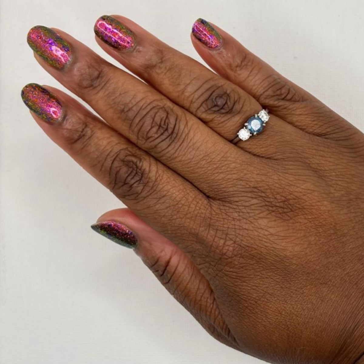 A close-up of a hand with metallic pink and green glitter nail polish, wearing a silver ring with blue and clear stones on the ring finger, set against a plain light background.