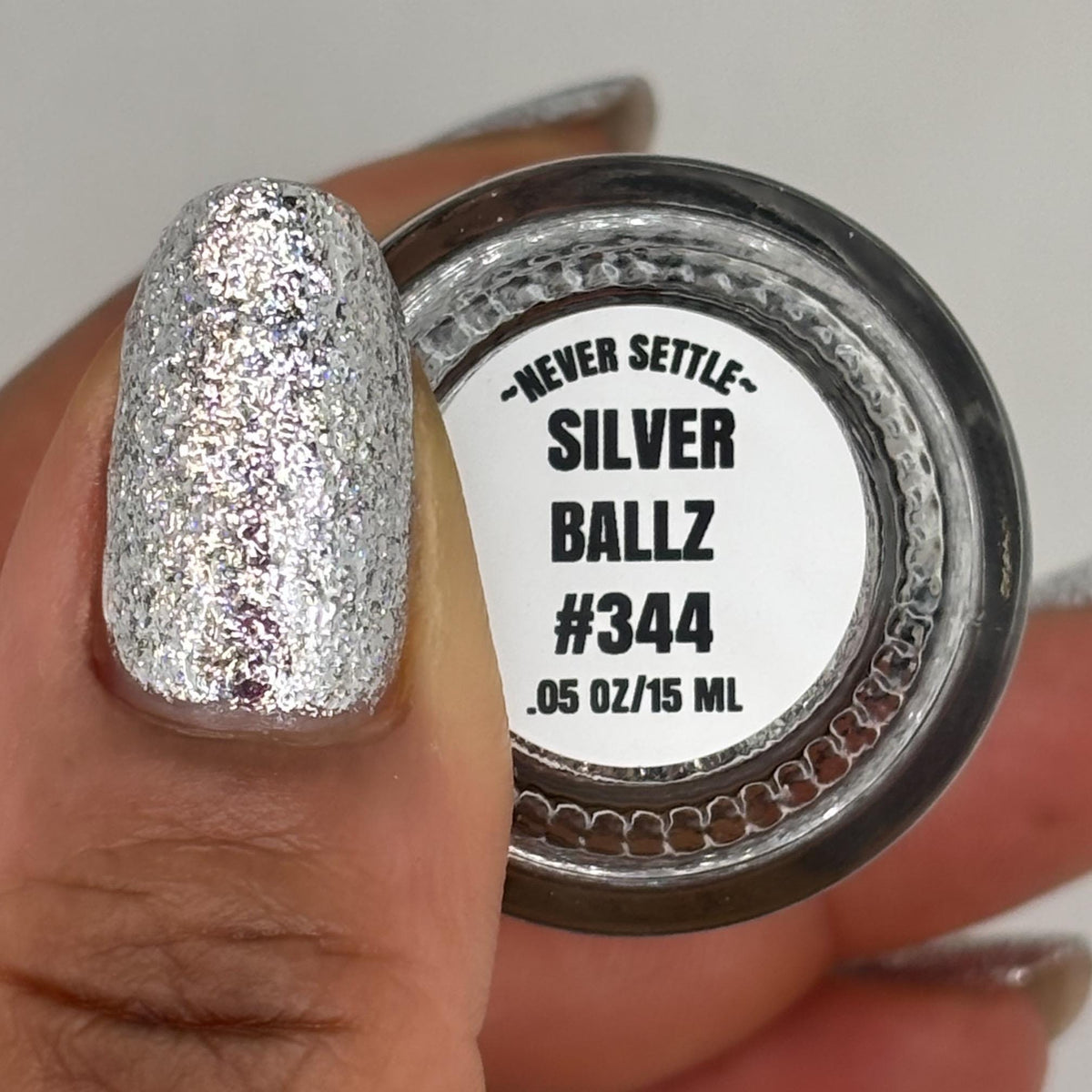 A close-up of a hand holding a bottle of glitter nail polish. The nail polish is applied in a sparkling silver shade to the fingernail, and the label on the container reads 'NEVER SETTLE SILVER BALLZ #344'.