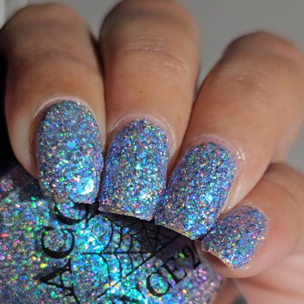 Close-up of a hand with nails painted in a dense holographic glitter nail polish, with the matching bottle partially visible in the background.