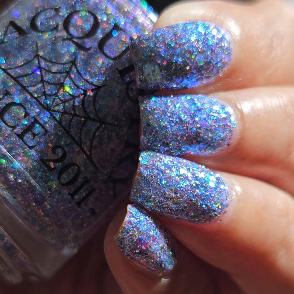 A close-up of a hand with nails painted in a sparkling holographic glitter polish, with the matching bottle partially visible on the left. The polish reflects blue, purple, and pink iridescent tones.
