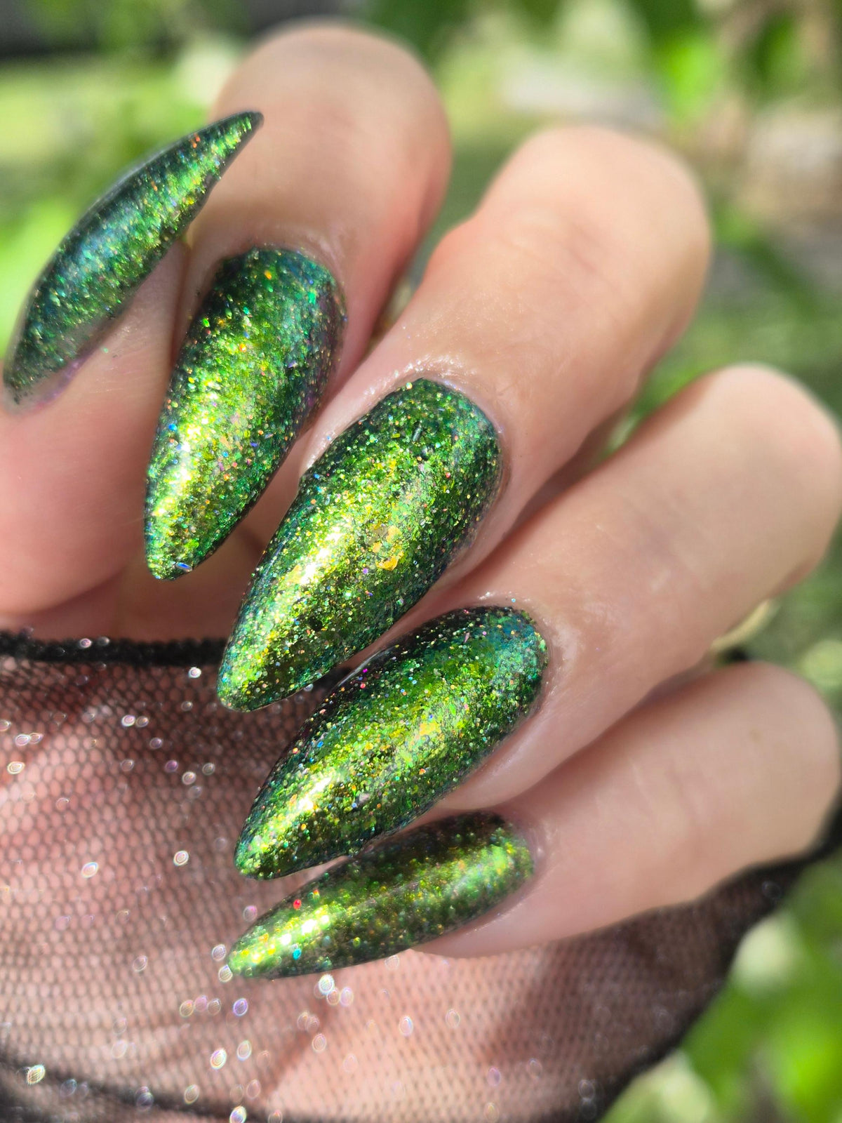 A close-up of a hand with long, stiletto-shaped nails painted in a sparkling metallic green holographic glitter nail polish, with a black lace glove visible at the wrist against a blurred green outdoor background.