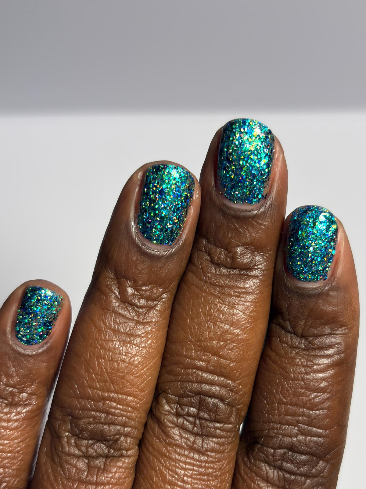 A close-up of a hand with fingernails painted in a sparkling teal and turquoise glitter polish against a light gray background.