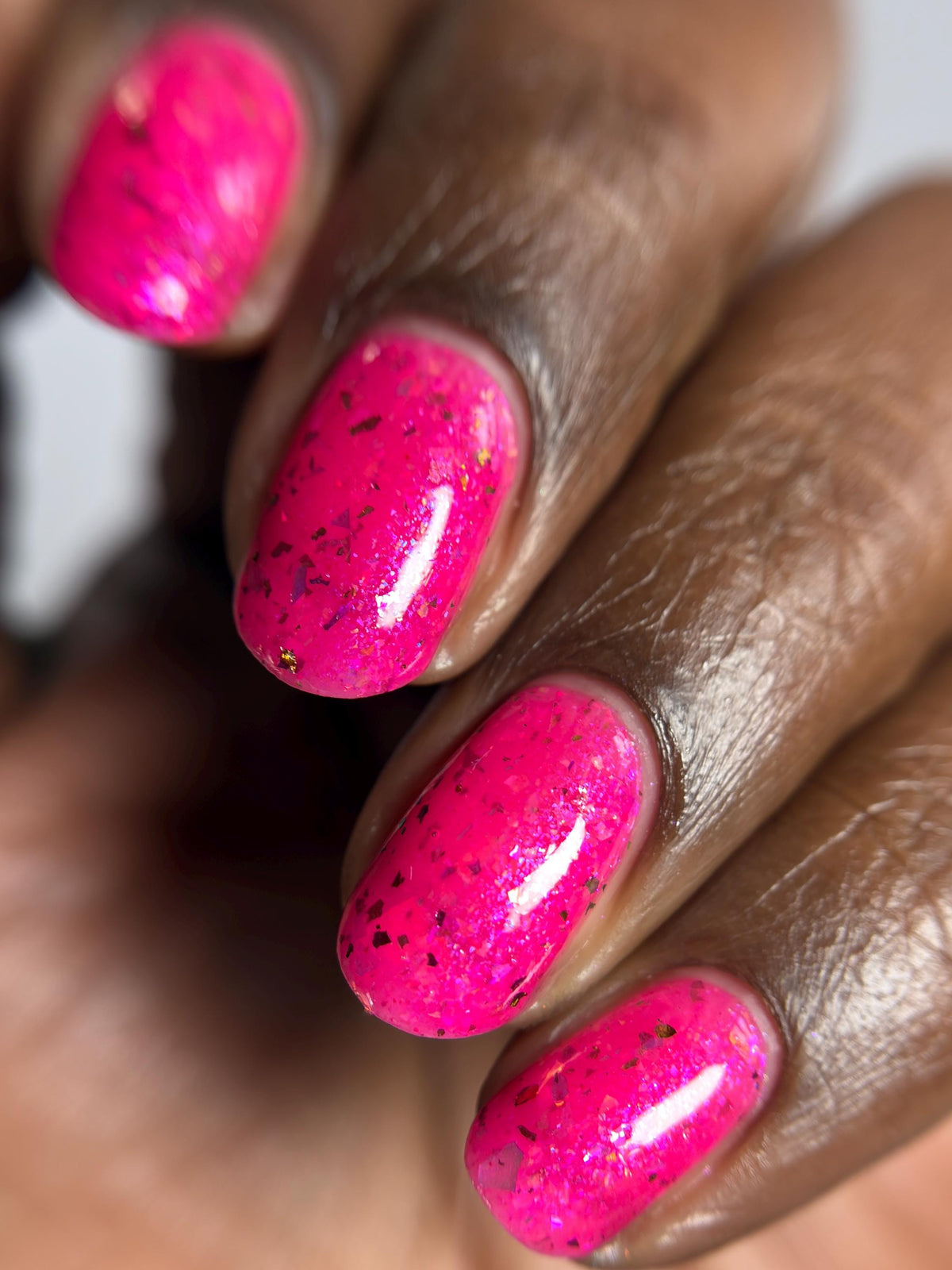 Close-up of a hand with dark skin with bright hot pink flake nail polish on short fingernails.