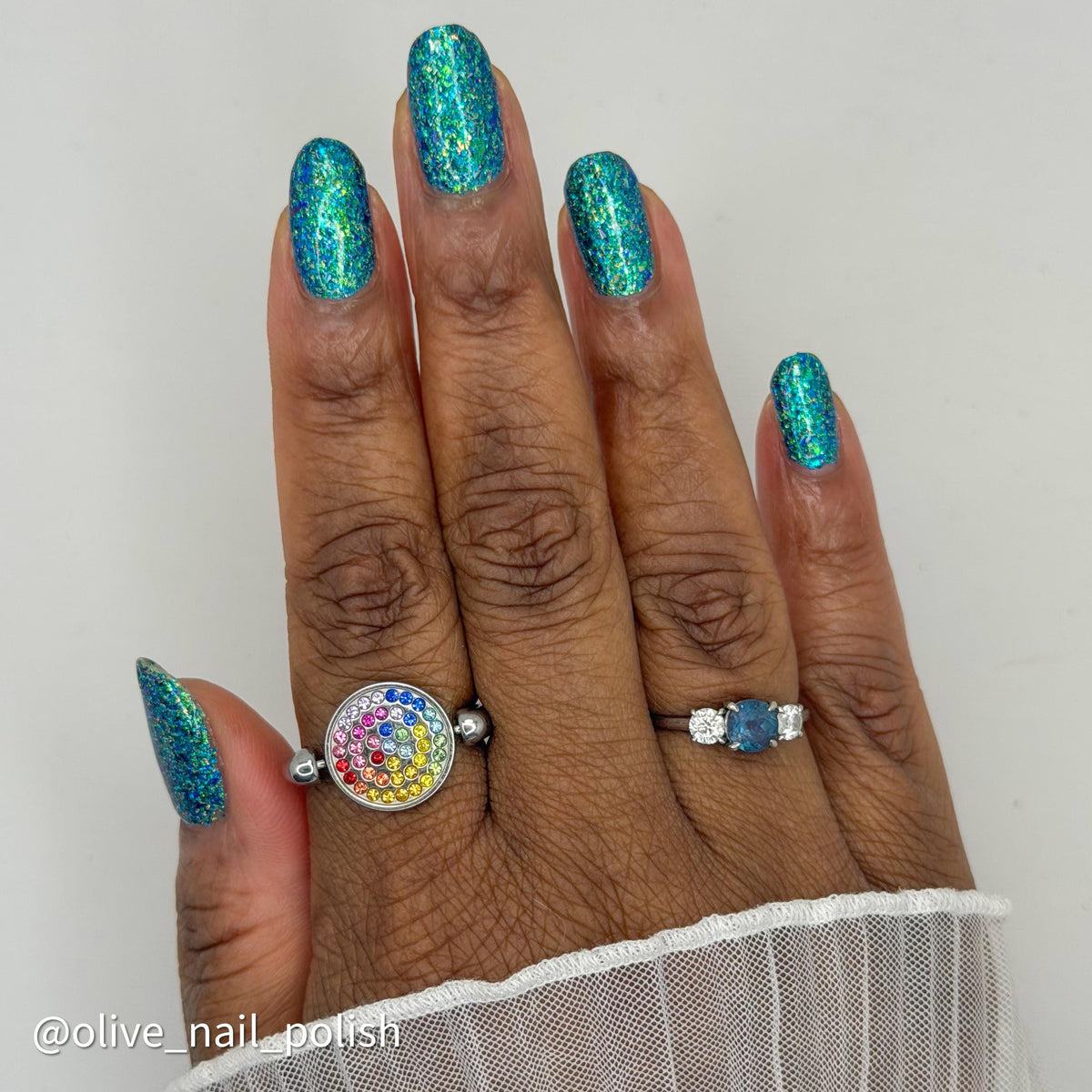 A close-up of a hand with medium-dark brown skin showcasing glittery teal blue nail polish, with colored rings