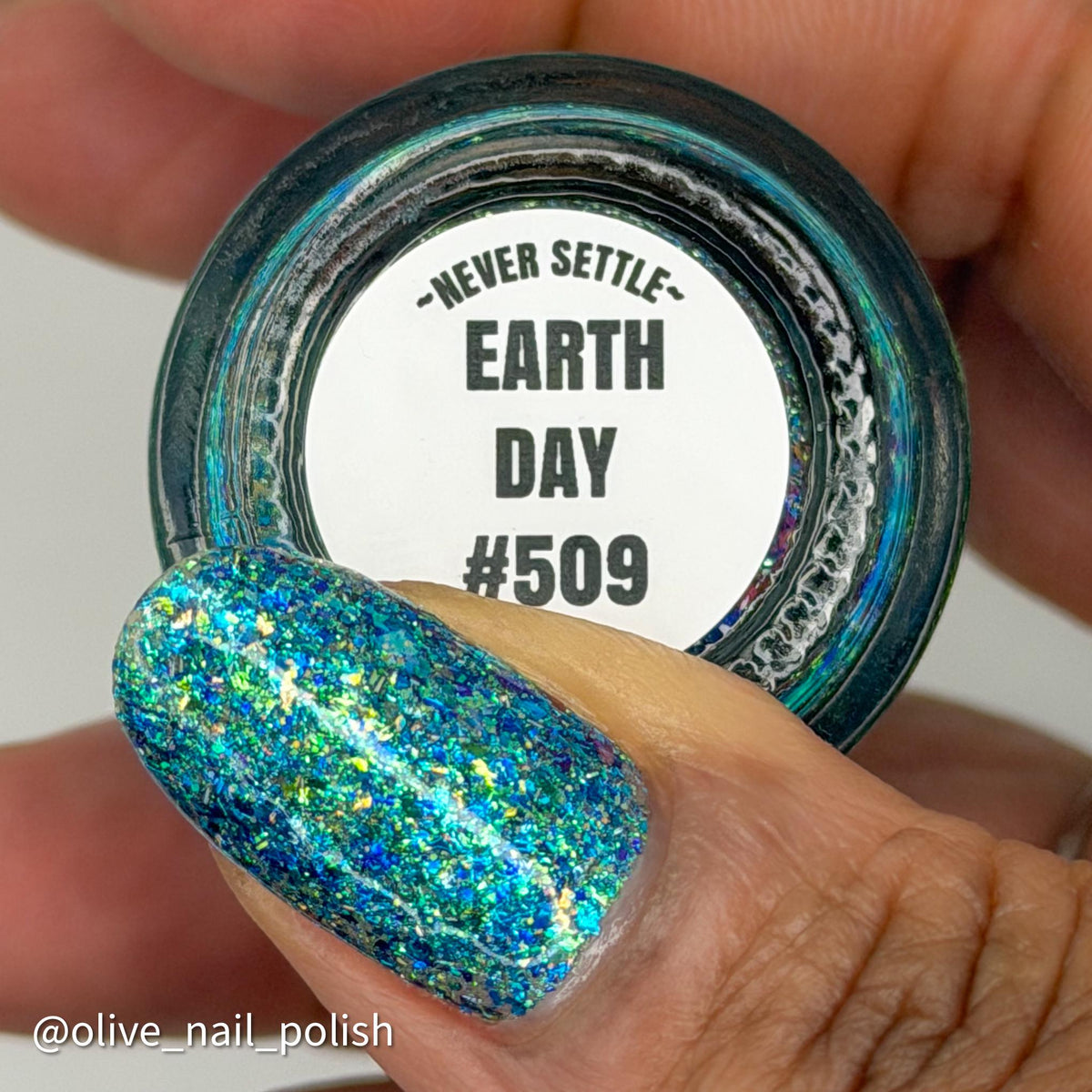A close-up of a hand holding a small round jar of nail polish with the bottom facing the camera. The nail polish being shown is a holographic glitter shade labeled 'NEVER SETTLE EARTH DAY #509' on the white label.