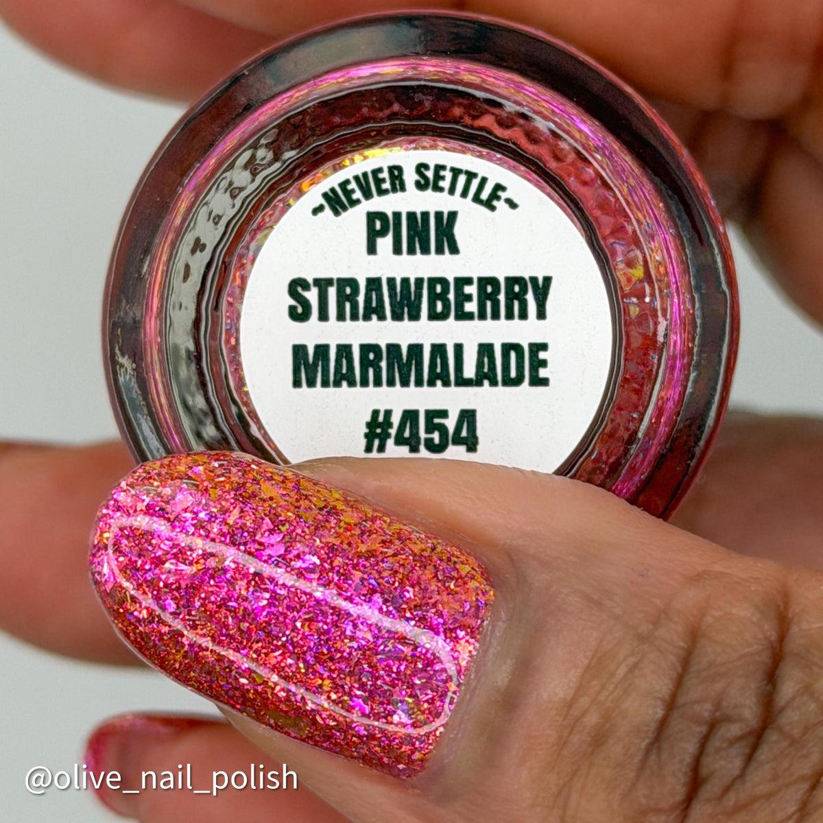 A close-up of a hand holding the bottom of a nail polish bottle with the label reading “Never Settle” and “Pink Strawberry Marmalade #454,” while a fingernail is shown painted in the same glittery pink polish. on a dark skin tone