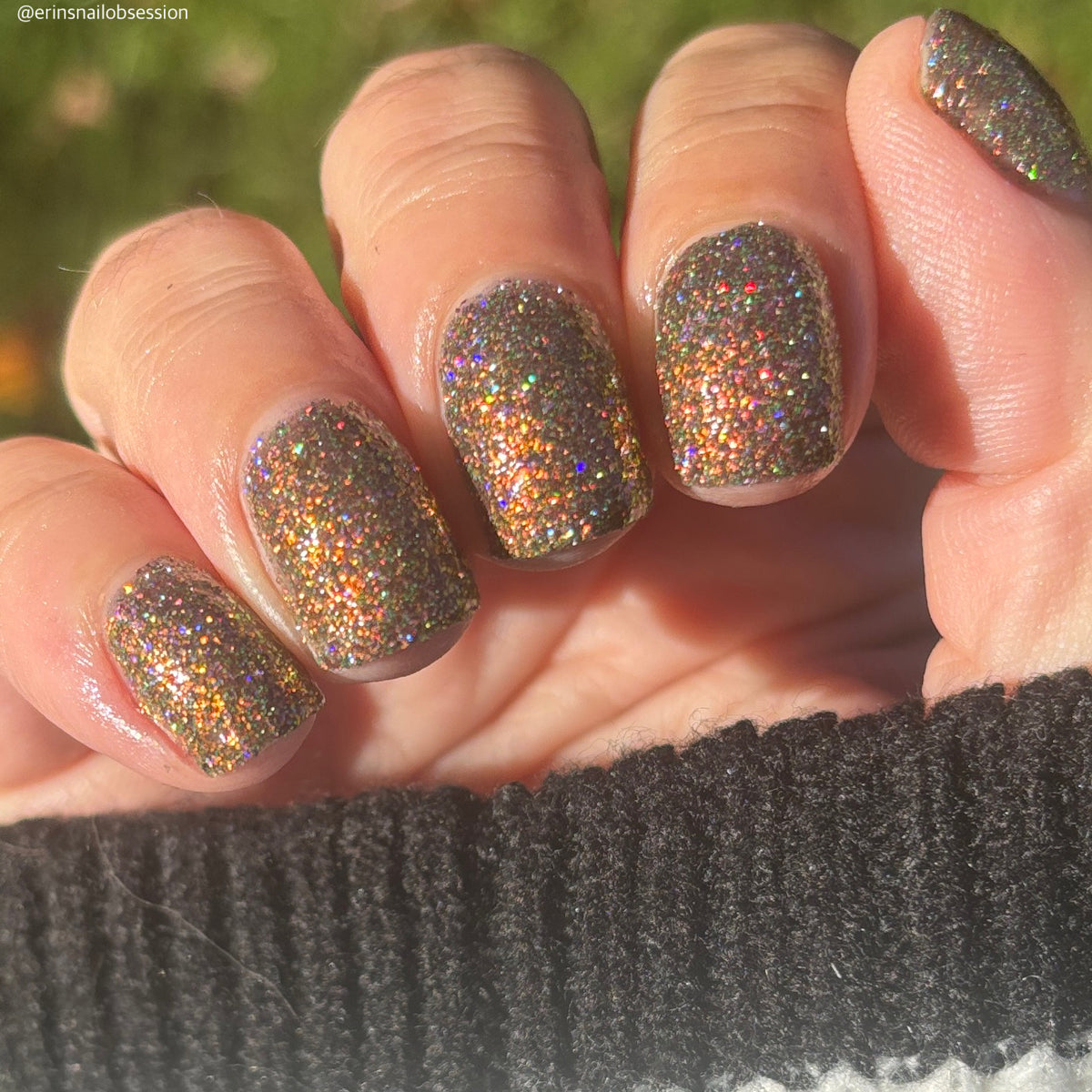nails painted with sparkly glittery polish showing pink, red, green, gold and orange glitters in a darkened base color with green reflective glitter
