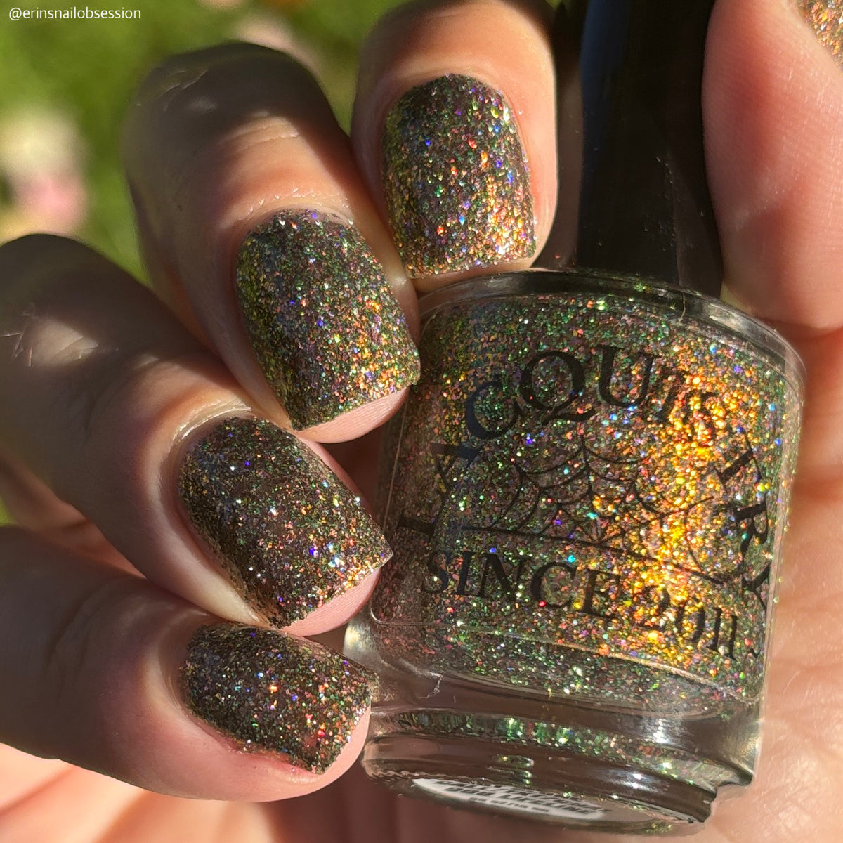 nails painted with sparkly glittery polish showing pink, red, green, gold and orange glitters in a darkened base color with green reflective glitter holding bottle that says Lacquistry