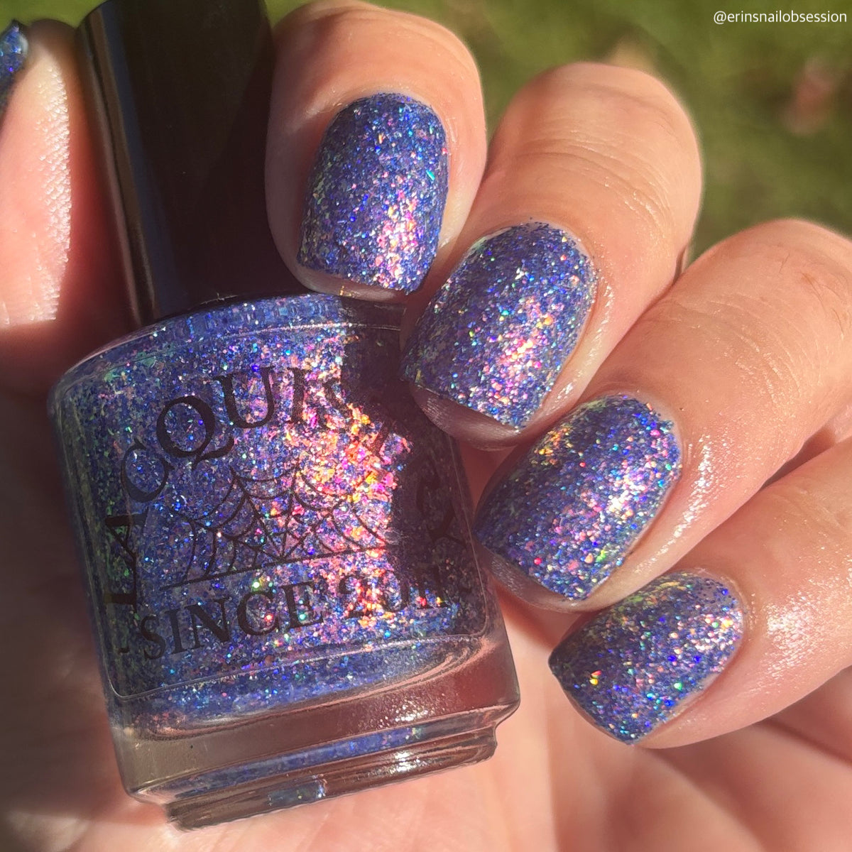A hand holding a bottle of glitter nail polish with matching glittered nails painted on, shown outdoors with blurred green grass in the background. The polish has a holographic blue-purple shimmer.