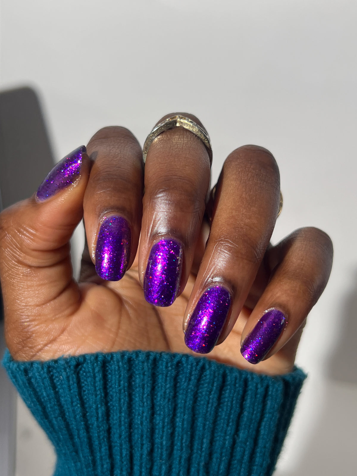 A close-up of a hand with manicured nails painted in a sparkling metallic purple polish, wearing a teal ribbed sweater cuff and a gold ring. dark skin tone