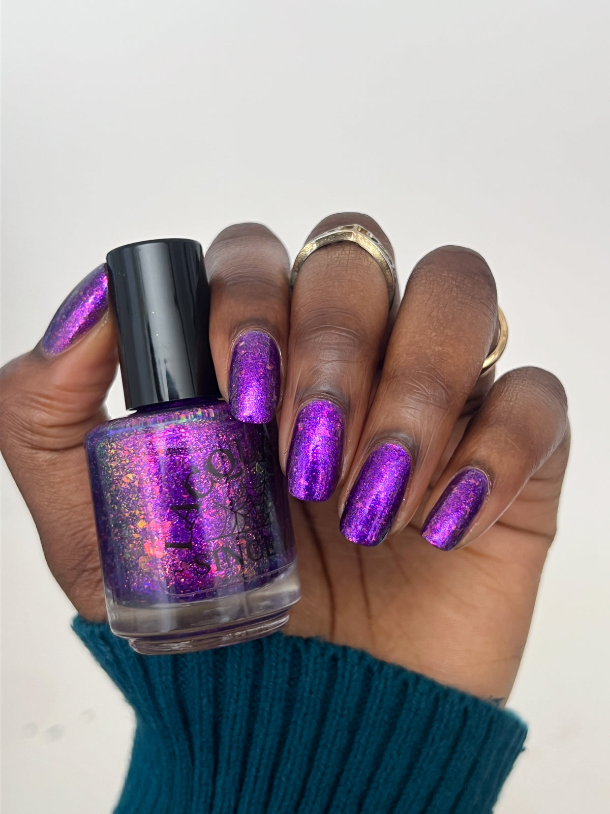 A hand with glittery purple and pink manicured nails holds a bottle of nail polish. The nail polish inside the clear bottle is a shimmering purple with pink and gold holographic flecks, and the bottle has a black cap.