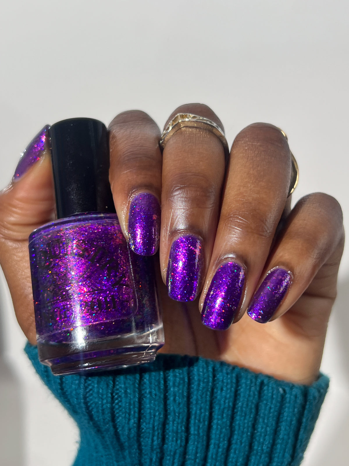 A hand holding a bottle of purple glitter nail polish that says "LACQUISTRY" next to matching purple glitter nail polish applied to the fingernails, with a teal knit sweater cuff visible against a plain light background.