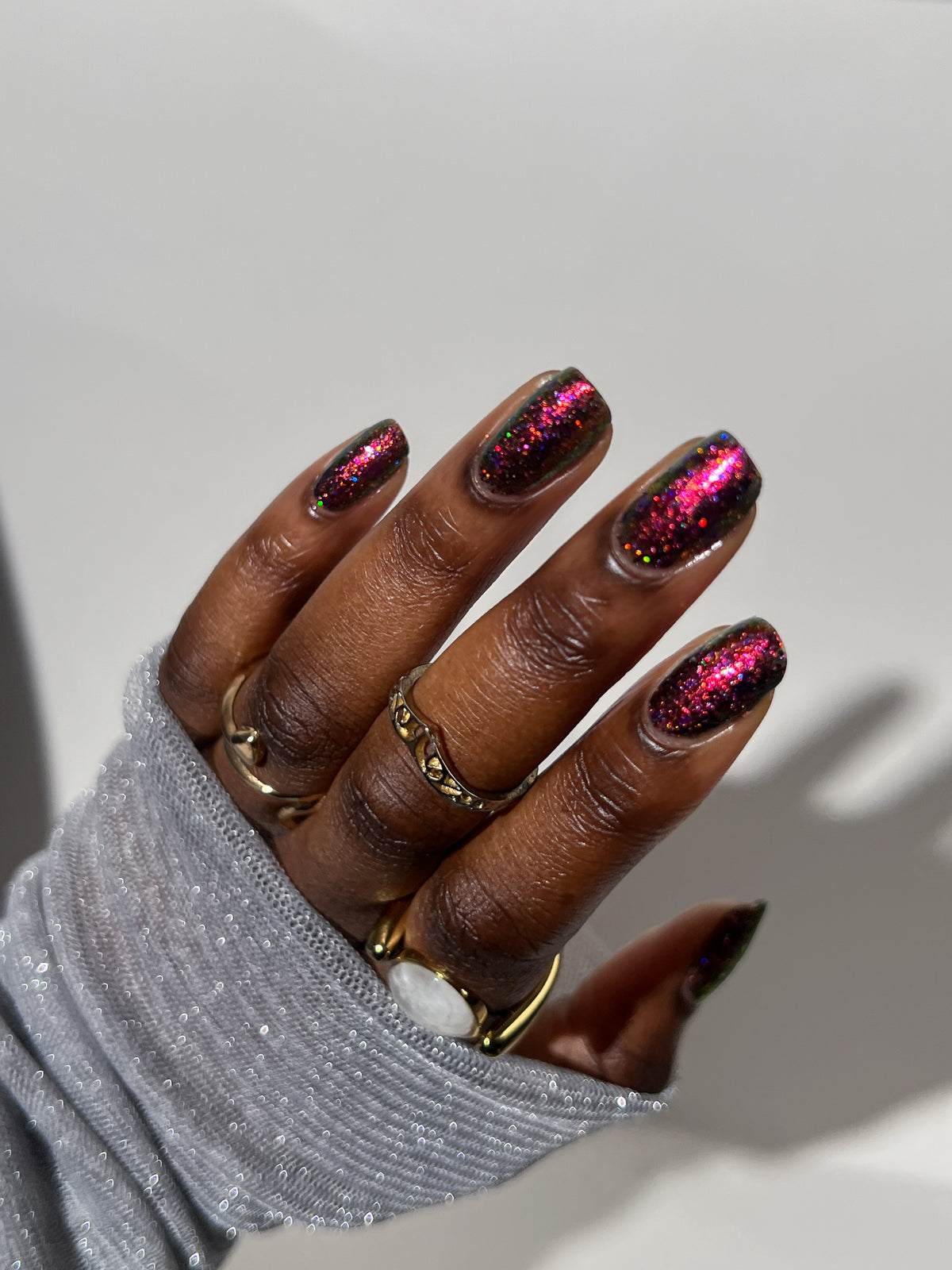 A close-up of a hand with manicured nails painted in a glittery dark pink and magenta gradient, wearing gold rings and a sparkly gray sleeve against a plain light background.